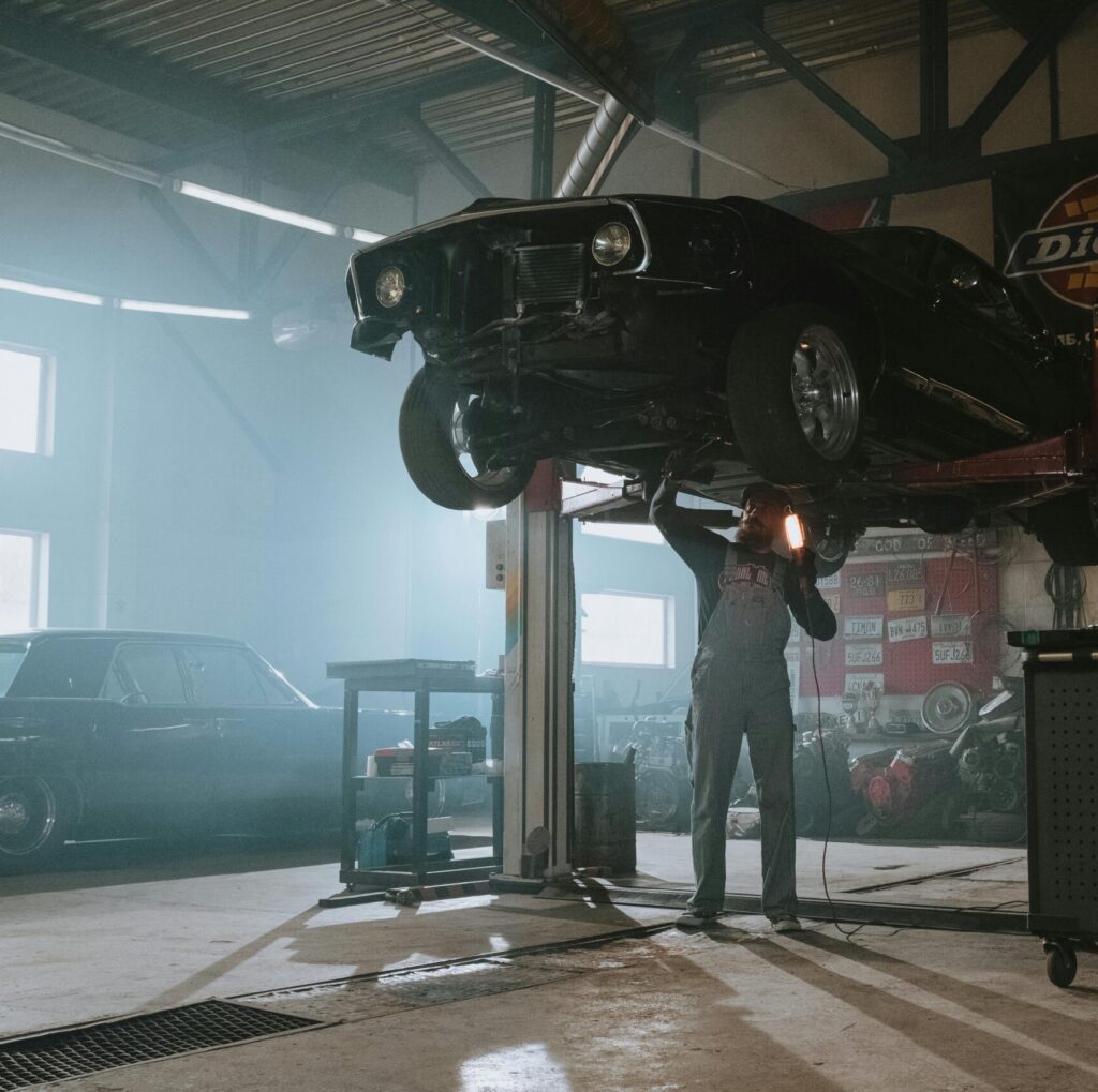 Male mechanic using a light to inspect a classic car lifted in an automotive garage.